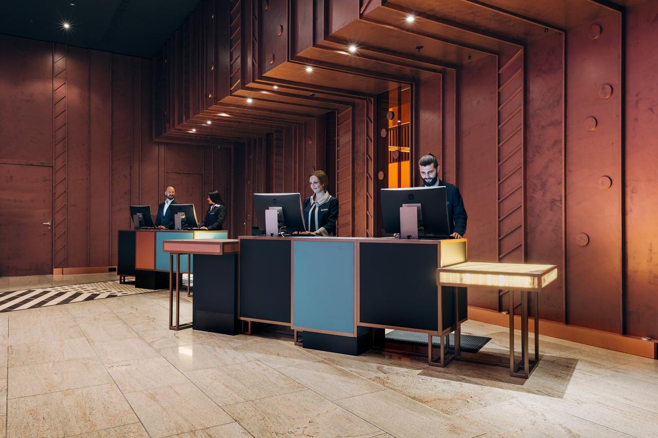 โรงแรมพูลแมน เบอลิน ชไวเซอร์ฮอฟ เบอร์ลิน ภายนอก รูปภาพ The image shows a modern hotel lobby reception area. There are several reception desks made of dark materials with illuminated accents. Behind the desks, three people are standing, likely staff members, engaged with computer screens. The lobby featur